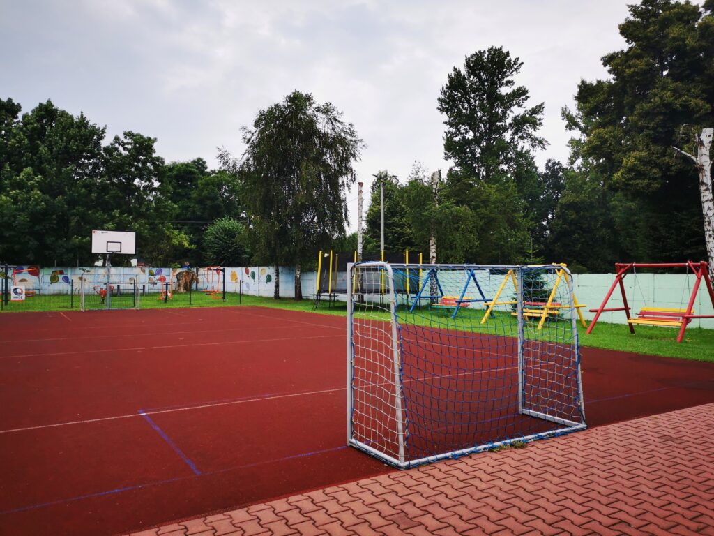 zdjęcie przedstawia boisko do gry w piłkę nożną. Oprócz dwóch bramek widać również kosz, siłownię, trampolinę oraz trzy huśtawki ogrodowe