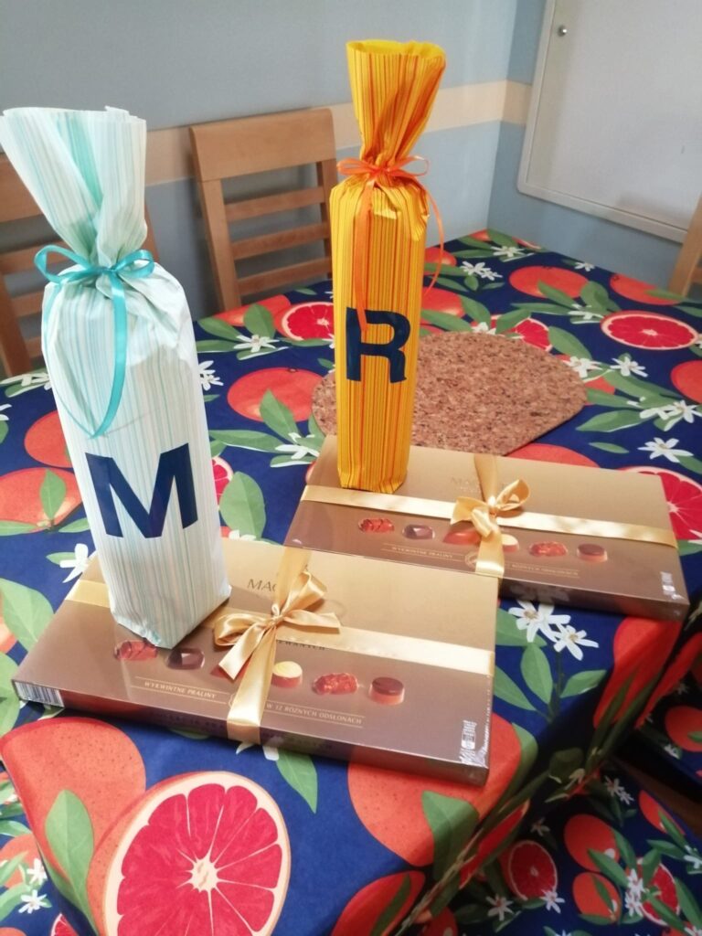 Na stole z kolorowym obrusem leżą dwie bombonierki oraz dwa pakunki prezentowe z literą M na jednym i R na drugim. 