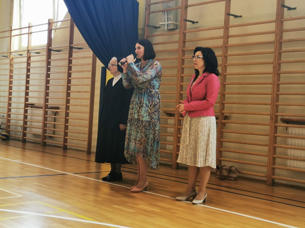 Hala sportowa. Trzy kobiety stoją, jedna z nich jest zakonnicą. Stojąca pośrodku kobieta przemawia do mikrofonu. 