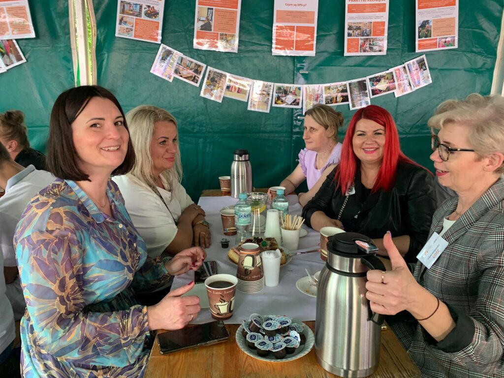 Grupa kobiet siedzi przy stole z poczęstunkiem i piją kawę. 