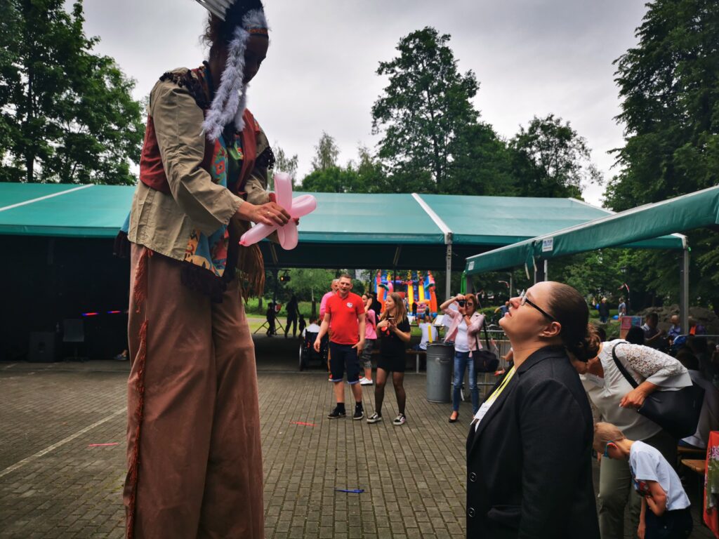 Kobieta stoi z podniesioną głową do góry. Przed nią kobieta na szczudłach przebrana za Indiankę robiąca figurkę z balonów do modelowania.  