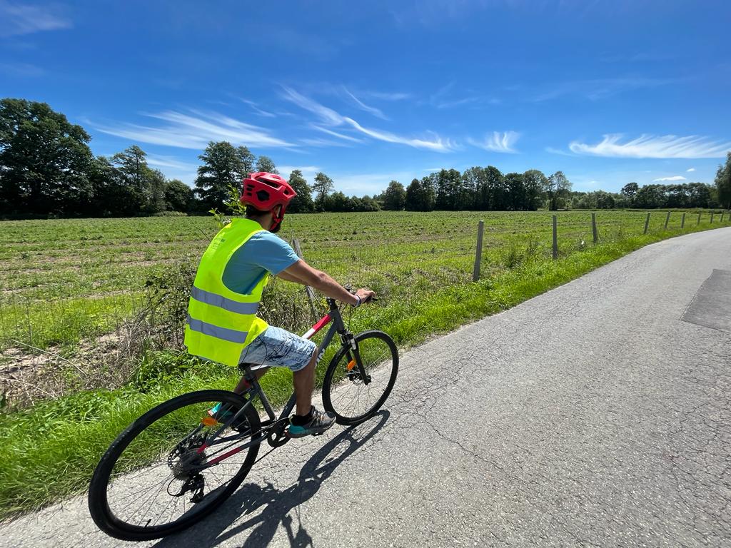 mężczyzna w czerwonym kasku i kamizelce odblaskowej jedzie na rowerze. Przed nim łąka, niebieskie niebo i droga. 