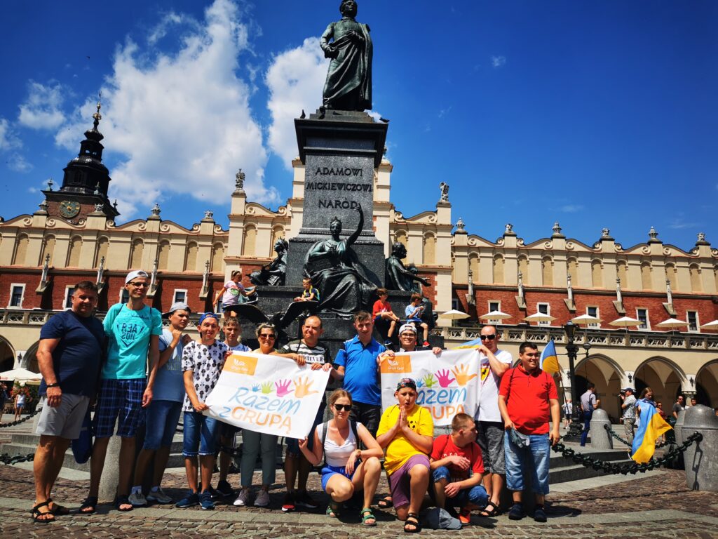 Grupa osób z plakatami "Razem z grupą" stoi przed posągiem Adama Mickiewicza na Krakowskim Rynku. 