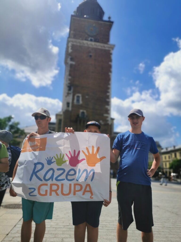 Troje chłopców trzyma plakat z napisem  "Razem z grupą". W tle ratusz krakowski. 