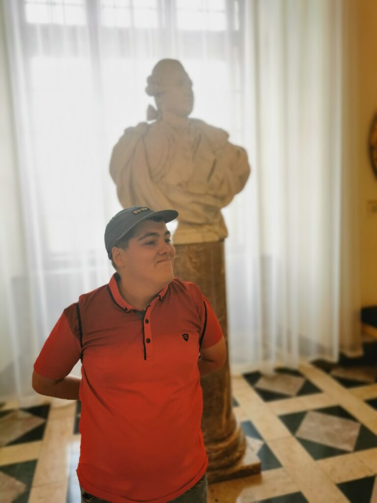 Chłopiec stoi przed popiersiem jakiegoś mężczyzny. Swoją postawą ciała naśladuje posąg. 
