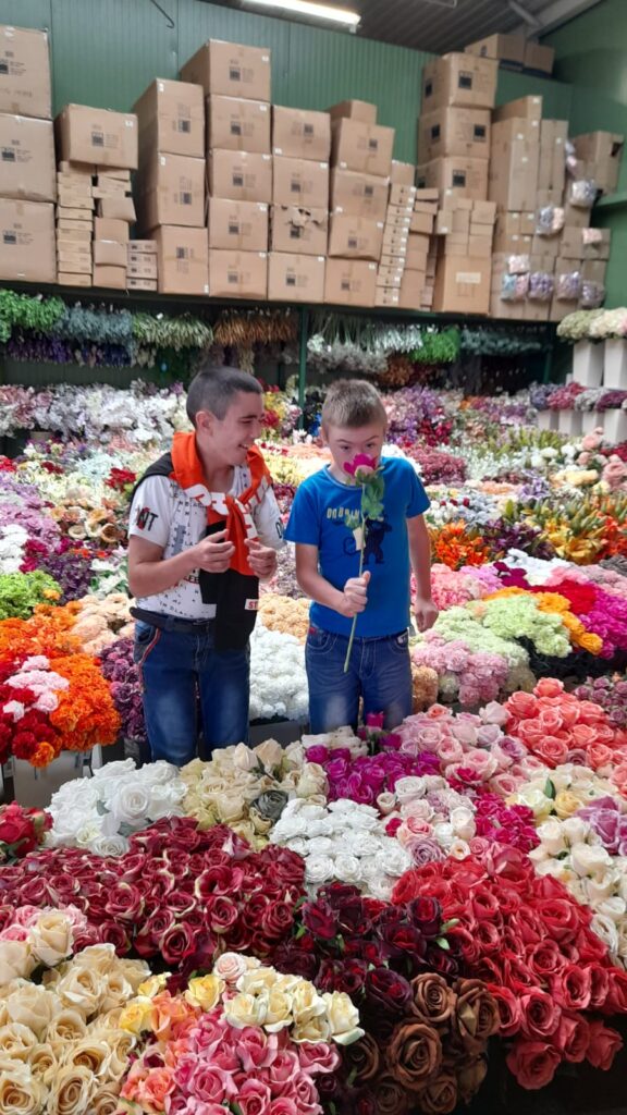 Dwoje chłopców stoi po środku pomieszczenia z kwiatami ciętymi. Jeden z nich się śmieje, drugi wącha kwiatek. 