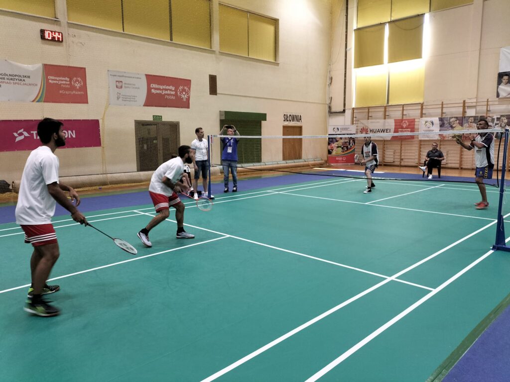 Wnętrze hali sportowej. Zielone boisko przedzielone siatką. Po obu stronach dwóch zawodników z paletkami do badmintona. 