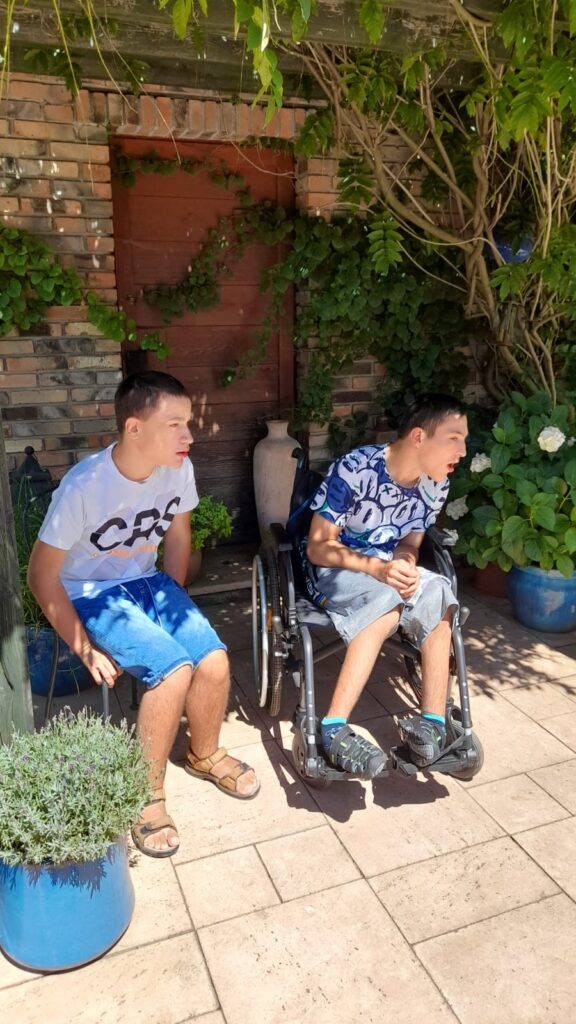 Dwoje chłopców (jeden siedzący na wózku) siedzą przed budynkiem. 
