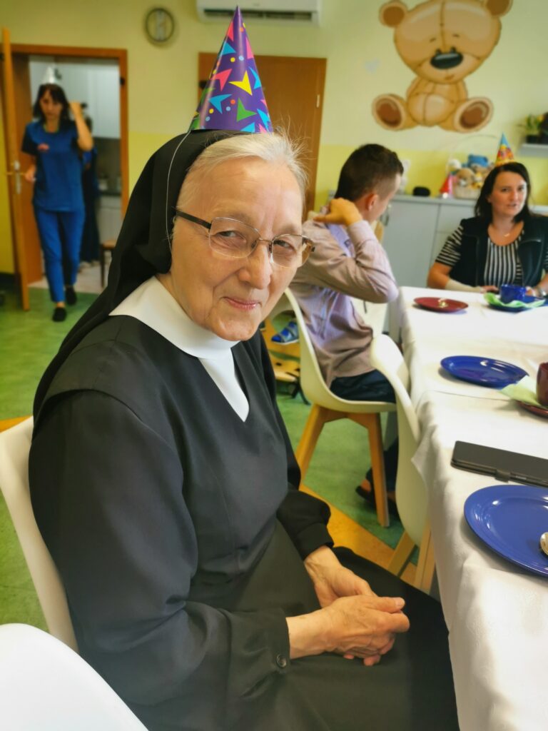 uśmiechnięta starsza zakonnica siedzi przy stole. Na głowie ma czapeczkę urodzinową. 