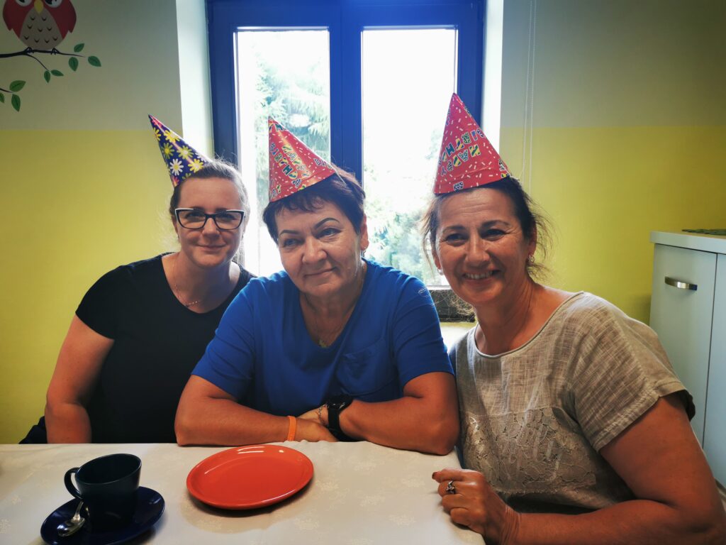 Trzy kobiety w kolorowych czapeczkach urodzinowych siedzą za stołem. 