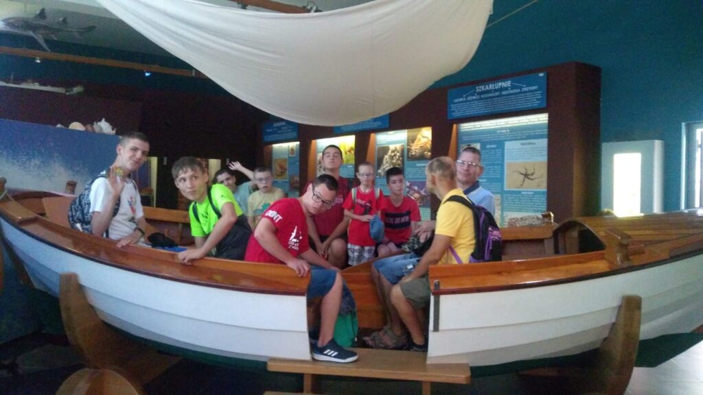 Grupa chłopaków w konstrukcji imitującej łódkę. Za nimi podświetlone gabloty z eksponatami. 