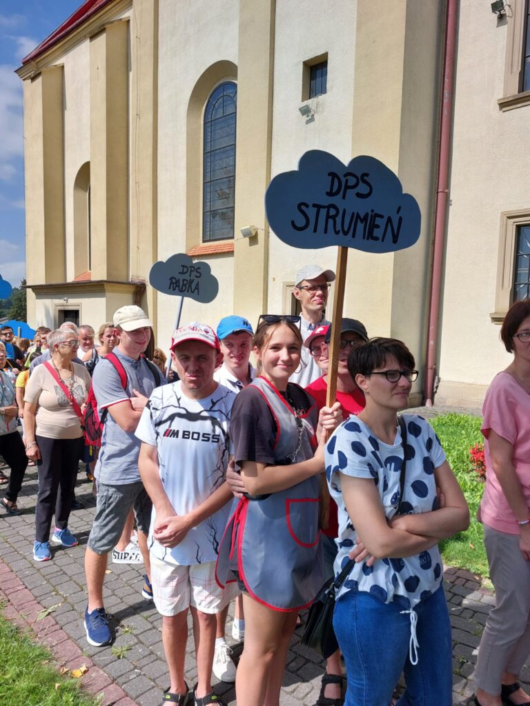 pochód wokół kościoła. Każda grupka ma tabliczkę z nazwą placówki. Na pierwszym planie kobieta trzyma tabliczkę "DPS Strumień" 