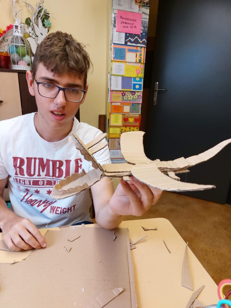 chłopiec przy stole w pracowni. W ręce prezentuje sklejone z kawałków kartonów latające stworzenie, które przypomina pterodaktyla. 