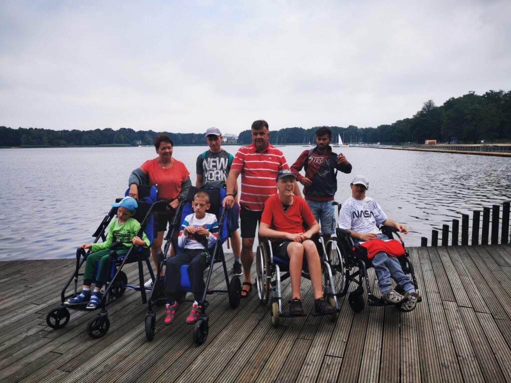 Grupa osób ( w tym 4 osoby na wózkach:) pozują do zdjęcia na tle jeziora. 