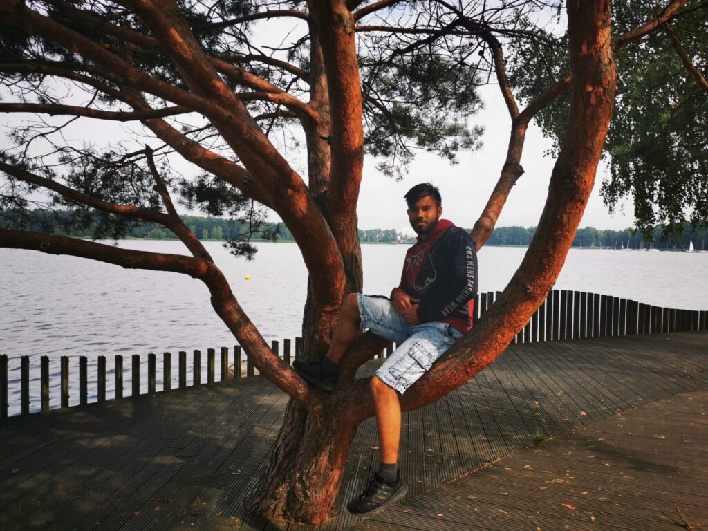 Młody mężczyzna siedzi na gałęzi na drzewie. Za nim jezioro.