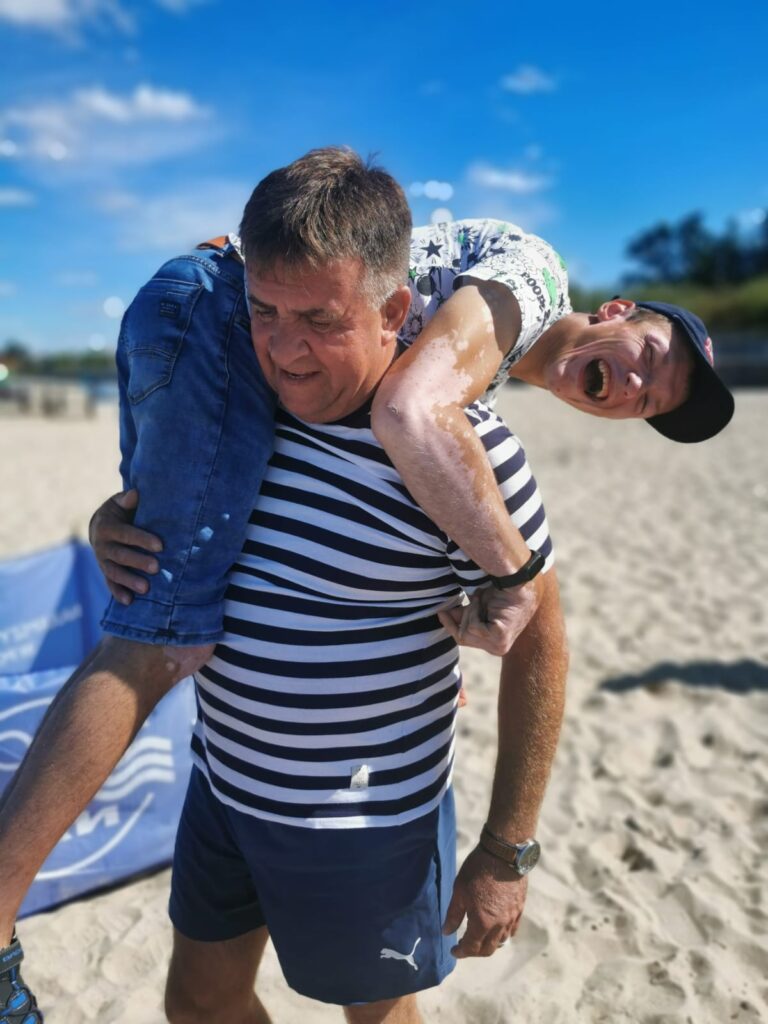 mężczyzna idący po plaży i niosący na plecach roześmianego chłopaka 