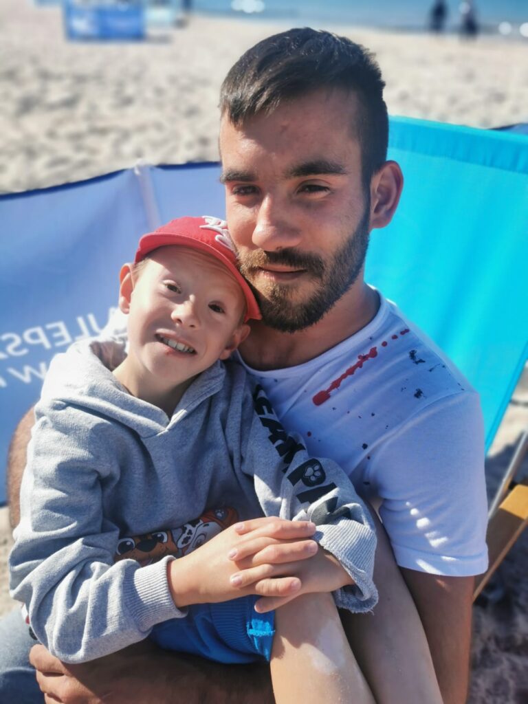 Chłopak siedzący na plaży i trzymający na kolanach młodszego kolegę 