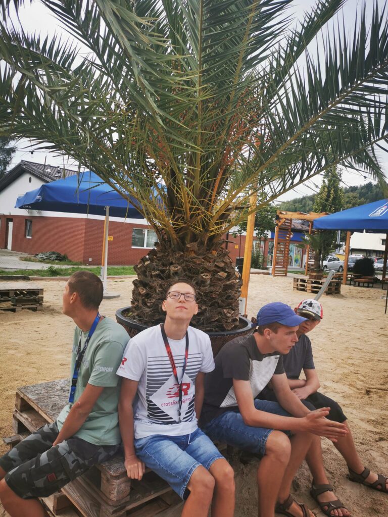 czworo chłopców siedzi na drewnianym podeście, wokół piasek a za nimi palma