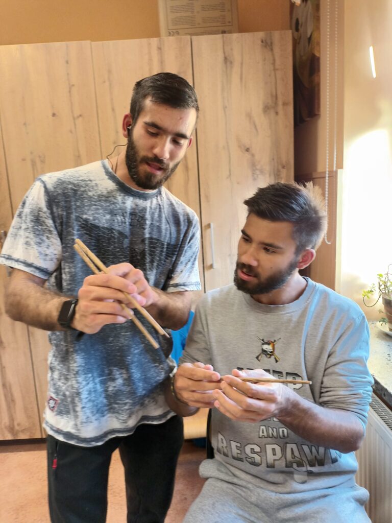 dwoje chłopaków trzyma w ręku pałeczki do sushi. Jeden pokazuje drugiemu jak chwycić przyrząd. 