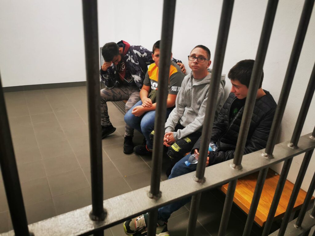 Grupa chłopców na ławce w celi. Na poierwszym tle kraty, 