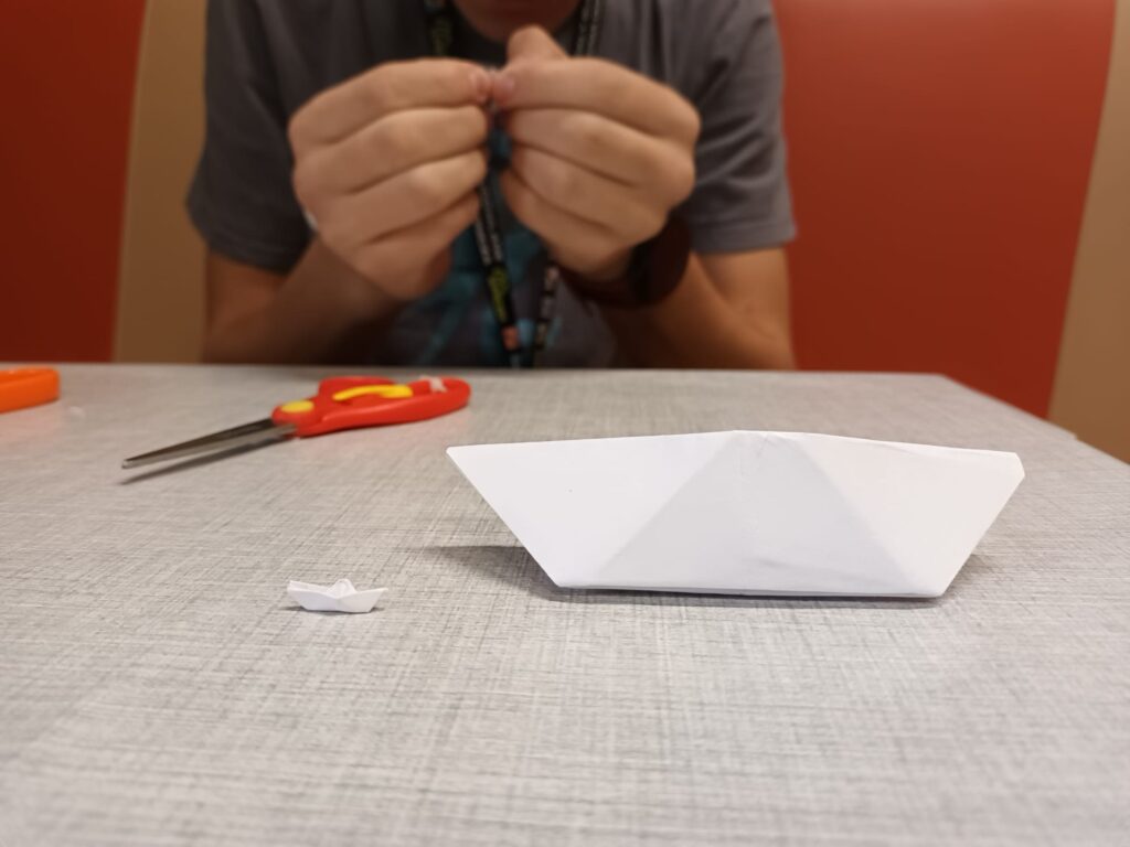 zdjęcie dwóch łódeczek z papieru na stole. Jedna z nich ma rozmiary zwykłej kartki, a druga jest miniaturowa ok. 5 milimetrowa. 