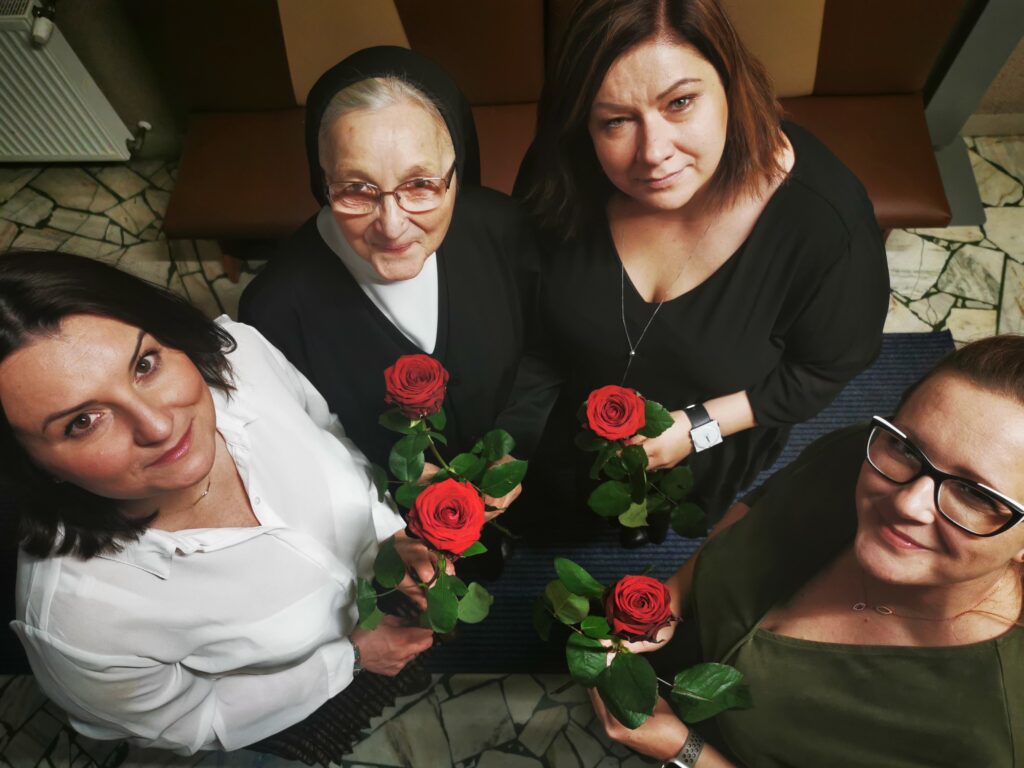 Cztery kobiety stojące w kółku. Patrzą w górę w obiektyw. Każda z nich ma wyciągniętą przed siebie różę 