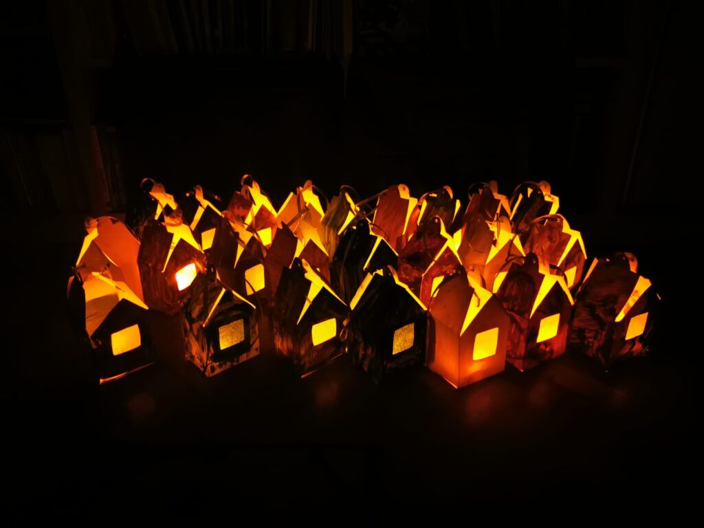 Na czarnym tle stoi duża ilość domków z papieru. W środku każdego z nich świeci lampka. 
