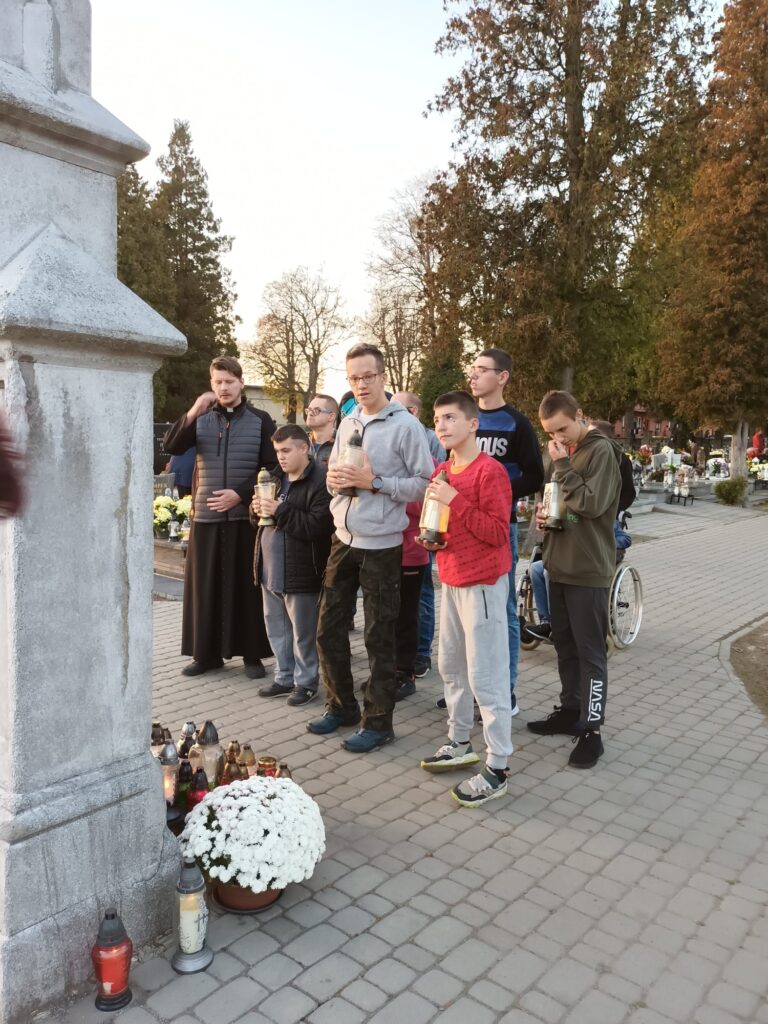 grupa osób pod krzyżem na cmentarzu. Każdy trzyma w dłoni znicze 