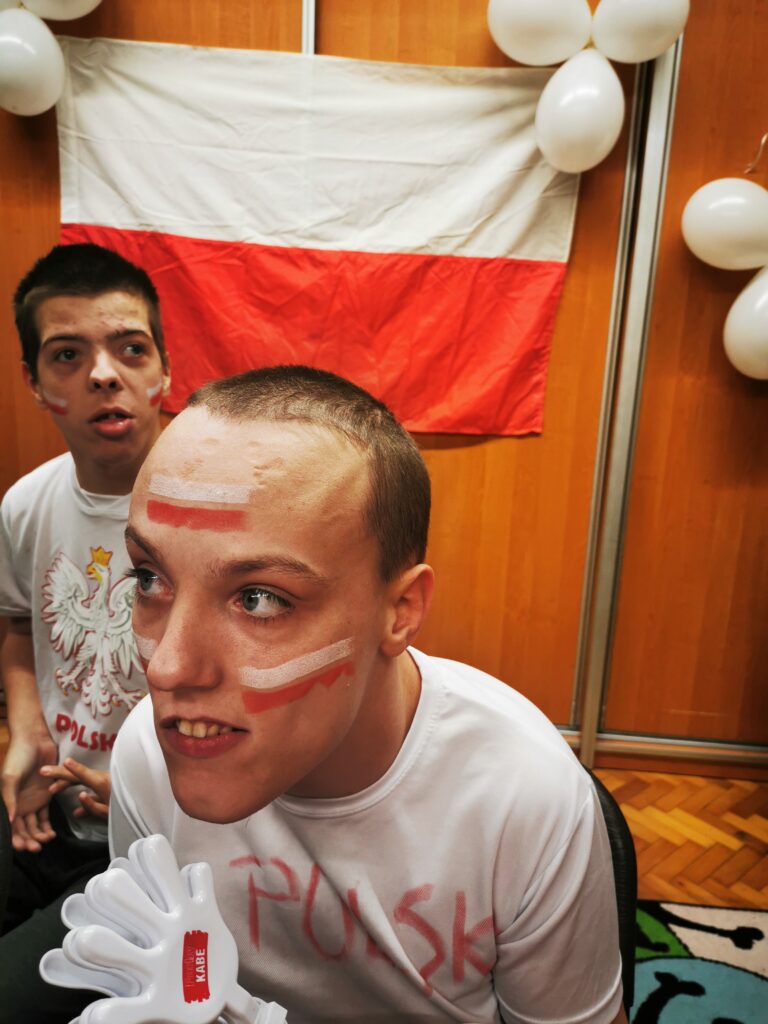 dwoje chłopców w białych koszulkach. Na twarzach mają namalowane polskie flagi. ZA nimi wiszą białe balony i biało-czerwona flaga 