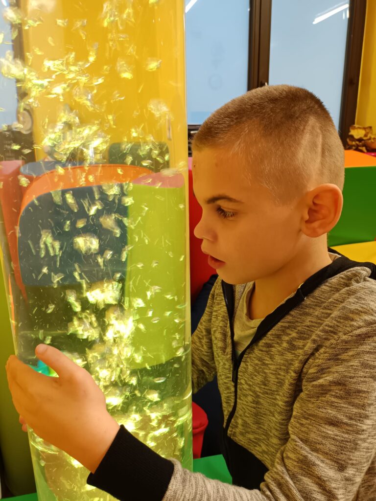 chłopiec przykłada czoło do dużej przezroczystej tuby, która napełniona jest wodą i wirują w niej bąbelki powietrza. Całość jest podświetlona na żółto. Chłopiec wpatruje się w bąbelki. 