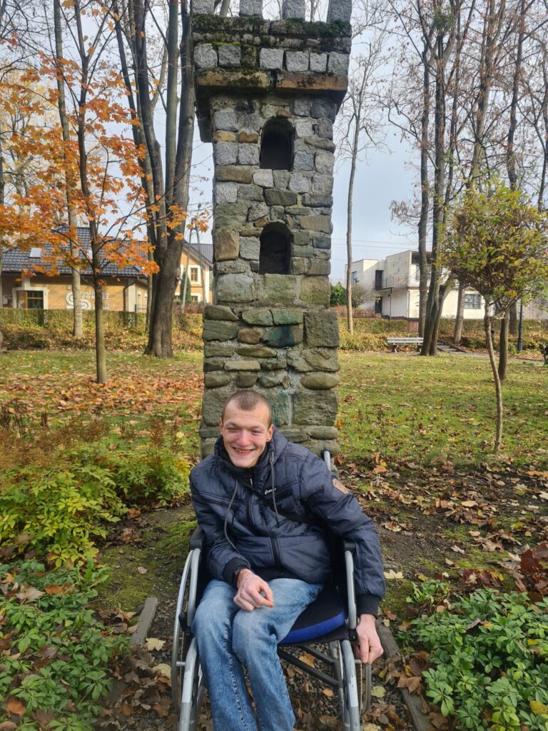 Mężczyzna na wózku uśmiecha się. 
Za nim kamienna miniatura wieży zamkowej. W tle drzewa 