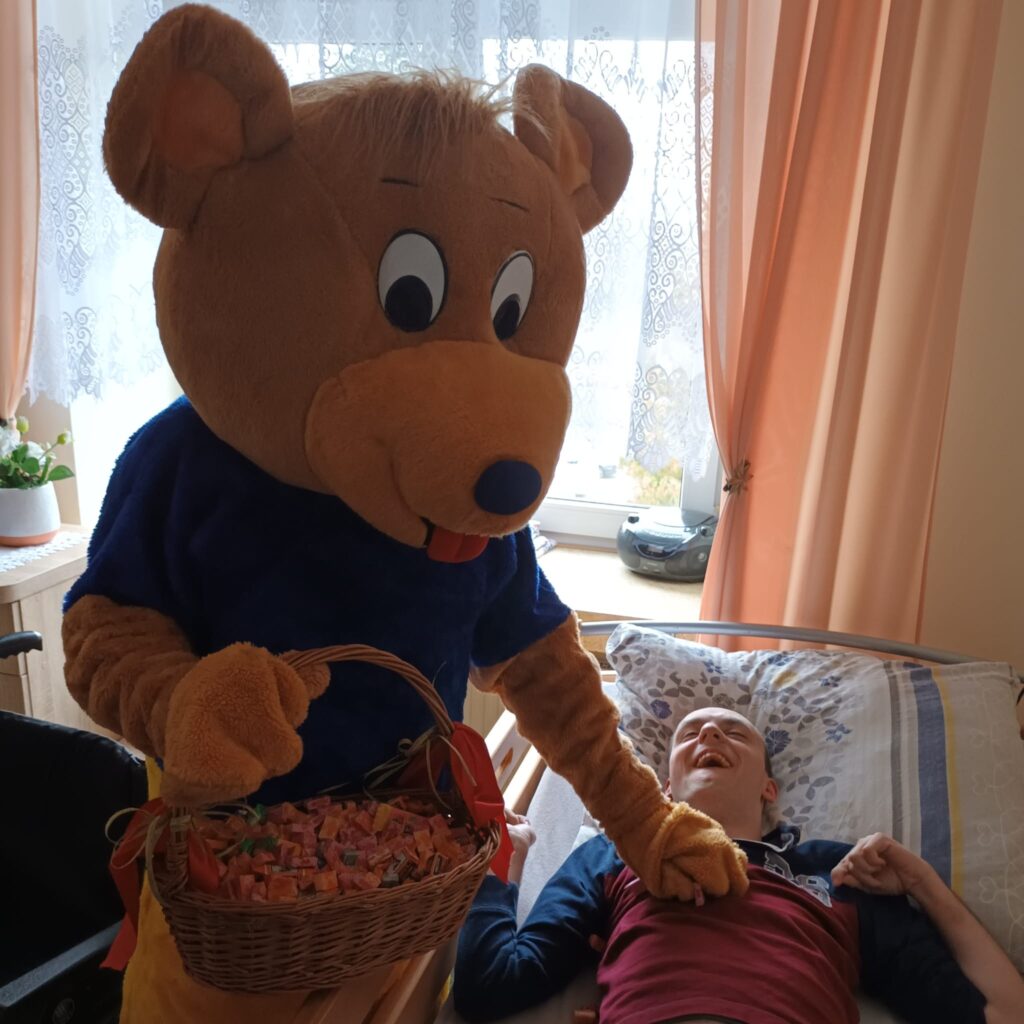 Osoba w przebraniu misia trzyma kosz z cukierkami i podaje rękę leżącemu na łóżku chłopakowi 