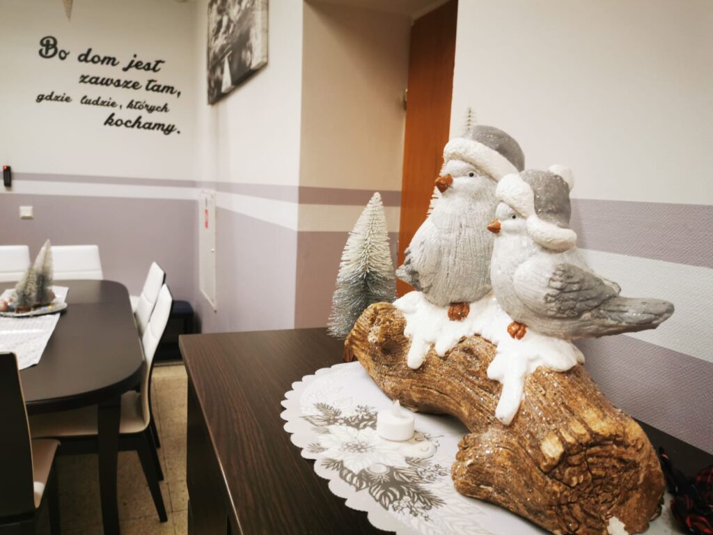 Świąteczna dekoracja: na półce położona jest serwetka, na której stoi figurka pieńka, na którym siedzą dwa ptaszki w zimowych czapkach. 