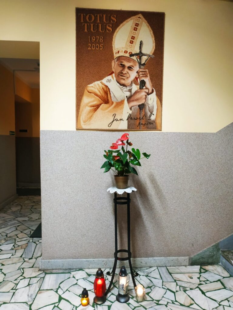 Ściana w pomieszczeniu, na której wisi obraz Jana Pawła II. Pod nim stoi kwietnik, a jeszcze niżej zapalone znicze. 