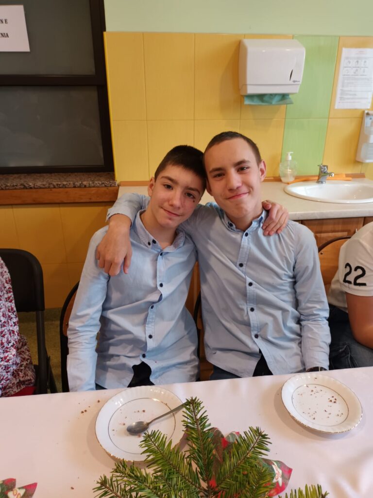 Dwóch ubranych w koszule chłopaków siedzi za stołem. Chłopcy obejmują się. 