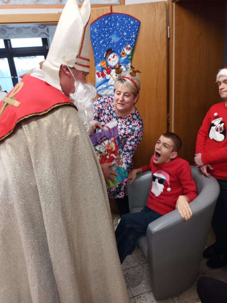 Szeroko uśmiechnięty chłopiec siedzi w fotelu. Na przeciw stoi osoba w kostiumie Św. Mikołaja i podaje mu prezent. 