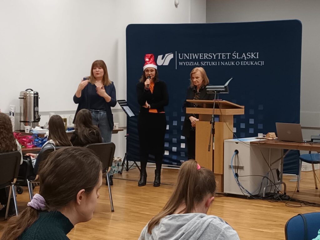 Dwie kobiety z mikrofonami na mównicy. Za nimi baner z napisem Uniwersytet Śląski. Obok stoi kobieta, która tłumaczy na język migowy. 