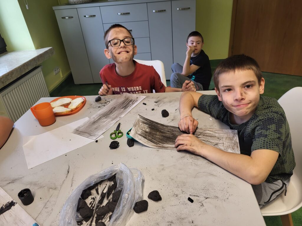 Grupa dzieci przy stole malująca na kartkach kawałkami węgla.  Twarze mają umorusane. 