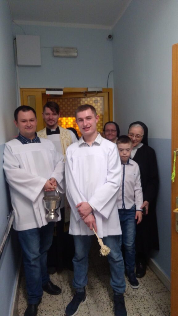Grupa osób stojąca w korytarzu: Ksiądz, dwie siostry zakonne, dziecko w białek koszuli oraz dwóch chłopaków w białych albach - jeden z nich trzyma metalowy pojemnik, a drugi kropidło 