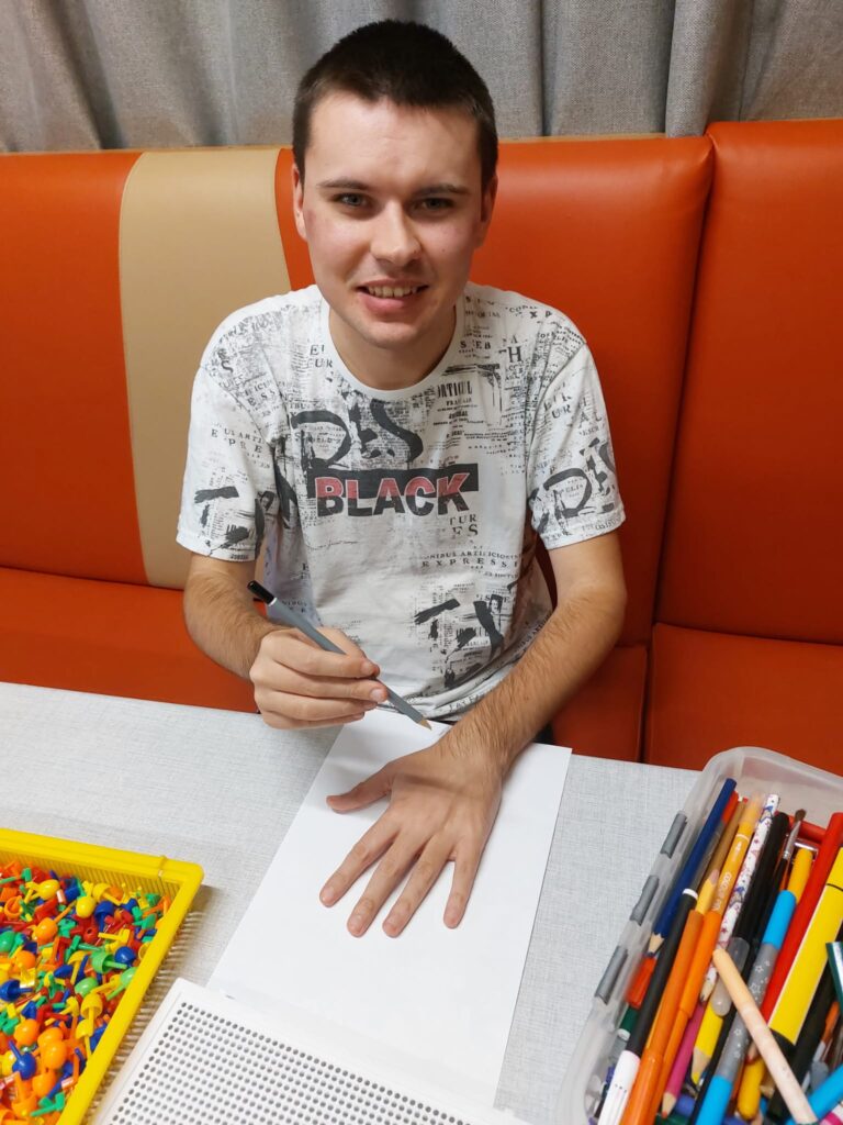 uśmiechnięty chłopak siedzi przy stole. W jednej ręce trzyma ołówek a drugą przykłada do leżącej przed nim kartki. 