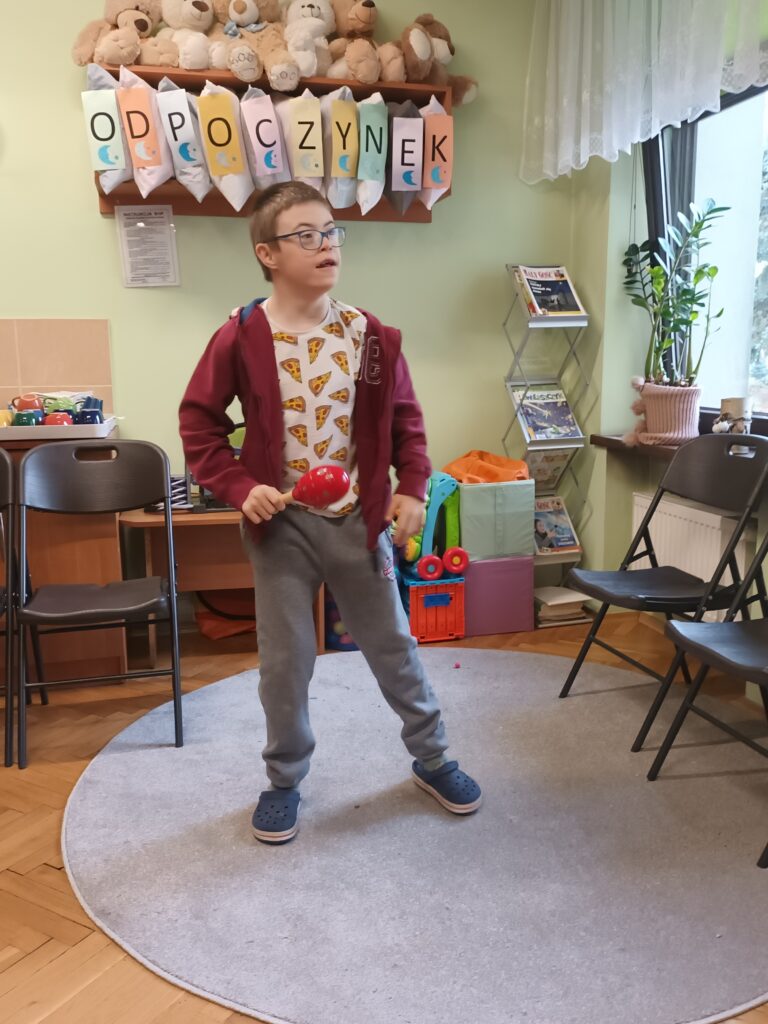 mały chłopiec z zespołem Downa stoi na dywanie. W ręce trzyma marakasy. 