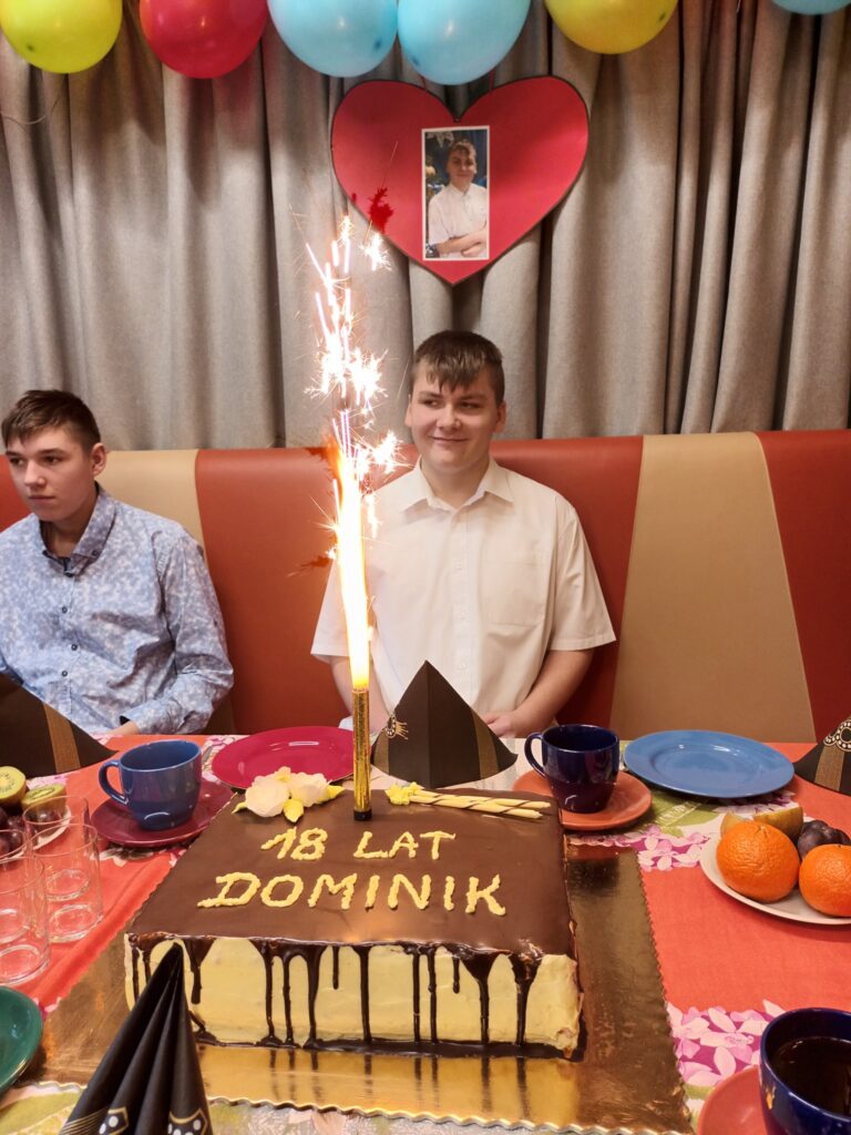 Płonąca raca wbita w tort z napisem "18 lat Dominik". Za tortem siedzi uśmiechnięty chłopak w białej koszuli. ZA nim na zasłonie wisi papierowe serce, na którym naklejone jest jego zdjęcie. 