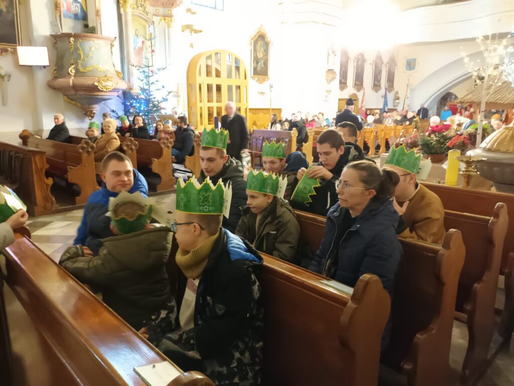 Osoby siedzące w ławkach w kościele. Na głowach mają zielone, papierowe korony. 