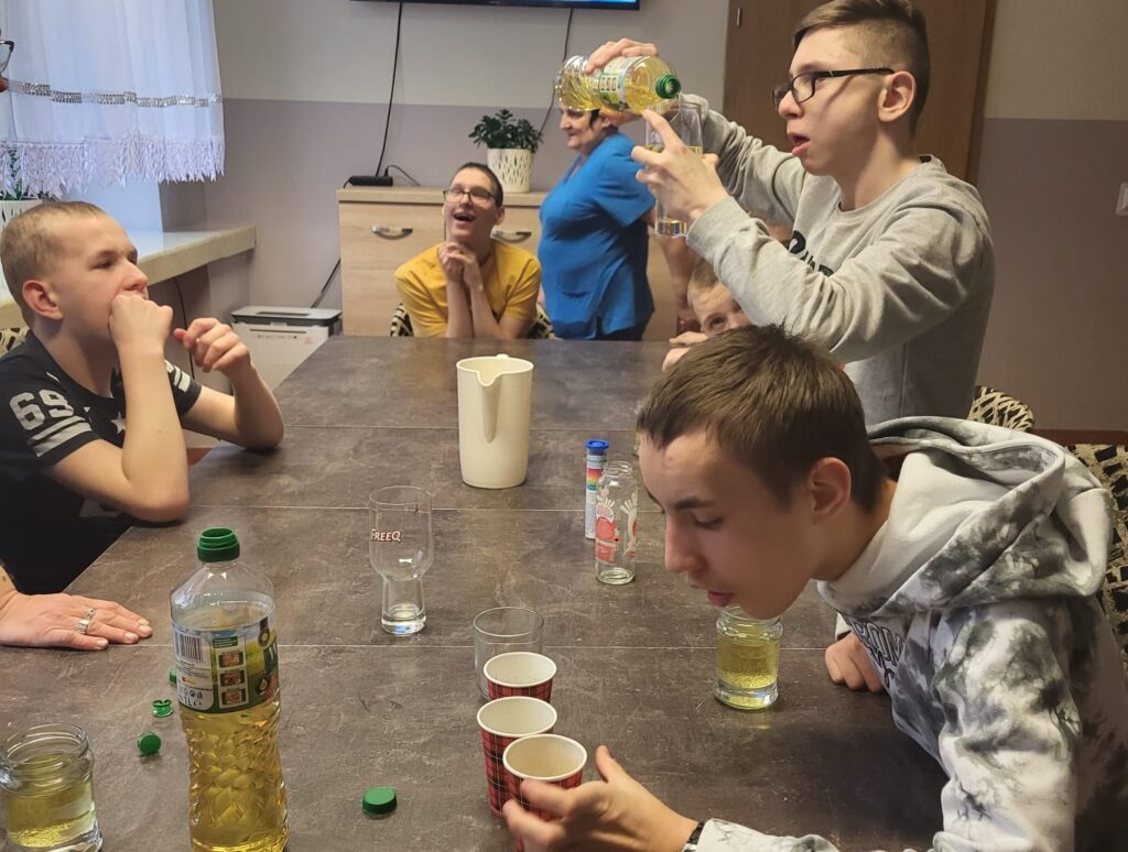 Grupa chłopaków wokół stołu. Na stole kubki i inne naczynia. Jeden z chłopaków leje do szklanki olej rzepakowy. 