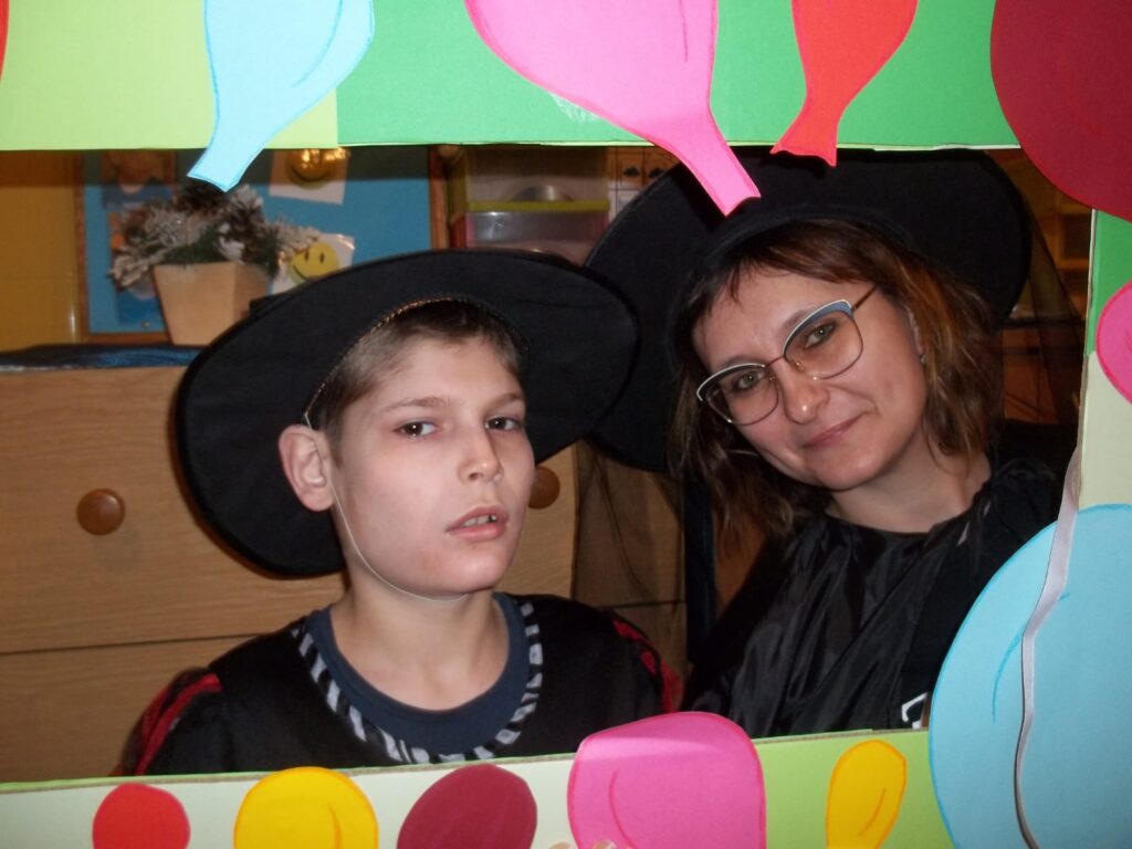 Kobieta i chłopiec w czarnych kapeluszach pozują do zdjęcia w tekturowej ramce  oklejonej kolorowymi, papierowymi balonami. 