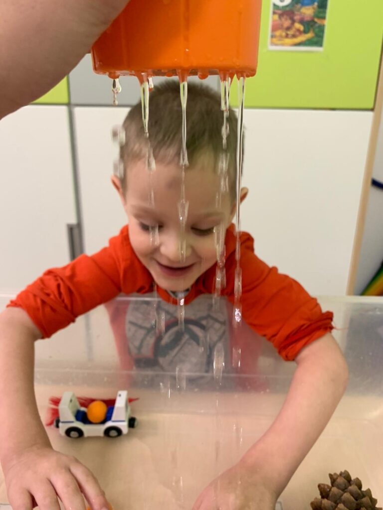 Uśmiechnięty chłopiec bawi się zabawkami w pojemniku z wodą. Z góry ktoś dolewa wodę przez coś przypominające sitko. 