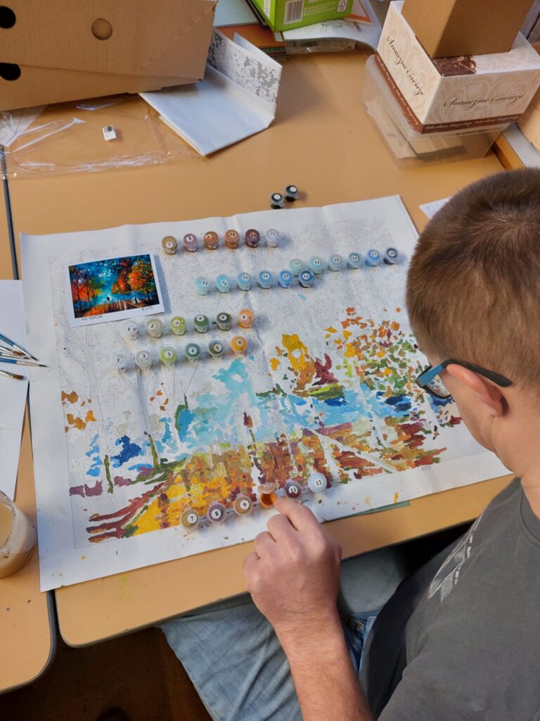 Chłopak malujący obrazek z dużą ilością małych elementów. Przed nim ustawione są ponumerowane małe farbki. 