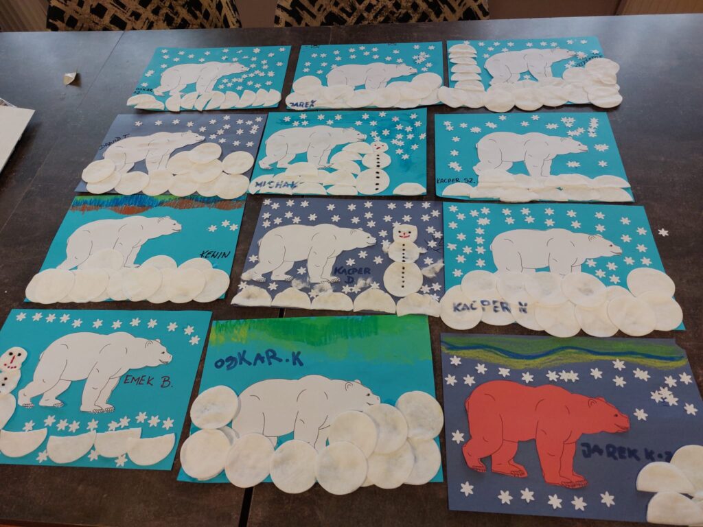 prace plastyczne ułożone na stole. Na niebieskich kartkach białe elementy wykonane z papieru i płatków kosmetycznych tworzące m.in. niedźwiedzie polarne, śnieżynki, bałwanki 