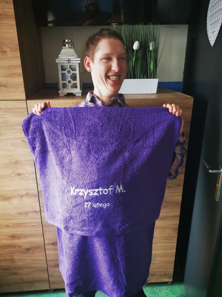 Uśmiechnięty chłopak trzyma w dłoniach fioletowy ręcznik z wyhaftowanym napisem "Krzysztof M. 27 lutego" 
