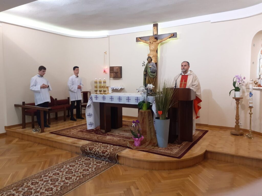 Mężczyzna ubrany w ornat stoi na mównicy przy ołtarzu. Za nim na ścianie duży krzyż. Obok przy ławce stoją dwaj ubrani na biało ministranci. 